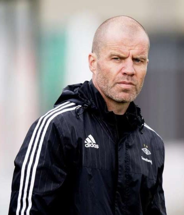 Ein kahlköpfiger Mann in einer schwarzen Jacke steht auf einem Fußballfeld, trägt eine marineblaue Jacke mit weißen Streifen und einem weißen Kragen und hat einen ernsten Gesichtsausdruck und die Hände vor der Brust verschränkt.