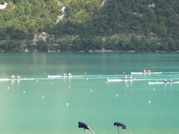 Gruppe von Menschen bei einem Ruderwettbewerb auf einem Gewässer mit Bäumen und einem Hügel im Hintergrund, Kleider über Stühle am Boden drapiert.