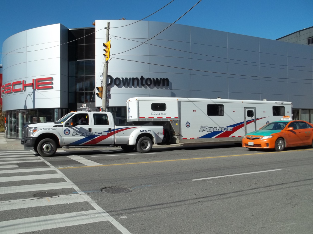 Polizeilkw vor einem Gebäude mit sichtbarem Text geparkt, Verkehrszeichen mit Masten und Kabeln im Vordergrund, Bäume und klarer blauer Himmel im Hintergrund.