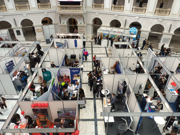 Eine große Gruppe von Menschen versammelte sich in einem Raum, der mit Ständen, Bannern, Tischen, Stühlen, Luftballons und einer Gebäudearchitektur mit Bögen, Säulen, Geländern und einem Bildschirm im Hintergrund gefüllt war.