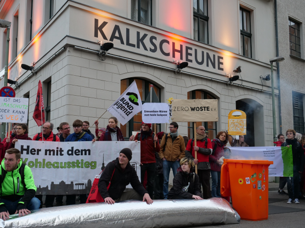 Eine Gruppe von Menschen mit Schildern und Plakaten vor einem Gebäude während einer Demonstration in Deutschland, mit zwei Menschen im Vordergrund und einem Müllcontainer auf der rechten Seite.