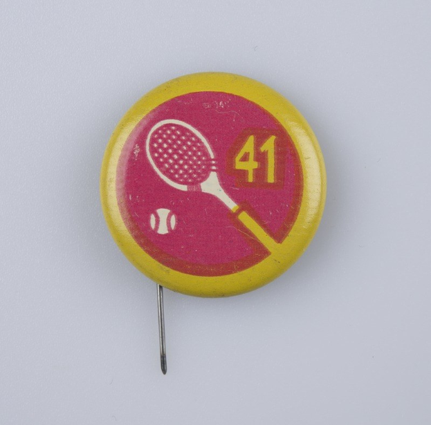 Gelb-rosa Abzeichen mit Tennisracket-Design, an einer weißen Oberfläche mit einer Nadel befestigt, mit Text und Zahlen.