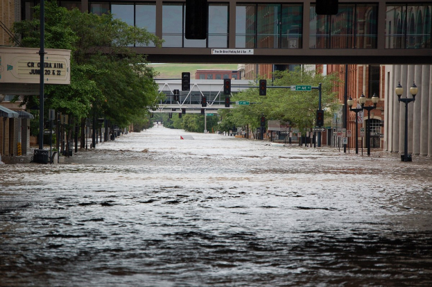 Überflutete Stadtstraße mit Wasser auf der Straße, Strommasten, Laternen, Verkehrszeichen, Bäumen, Gebäuden und einer Brücke im Hintergrund.