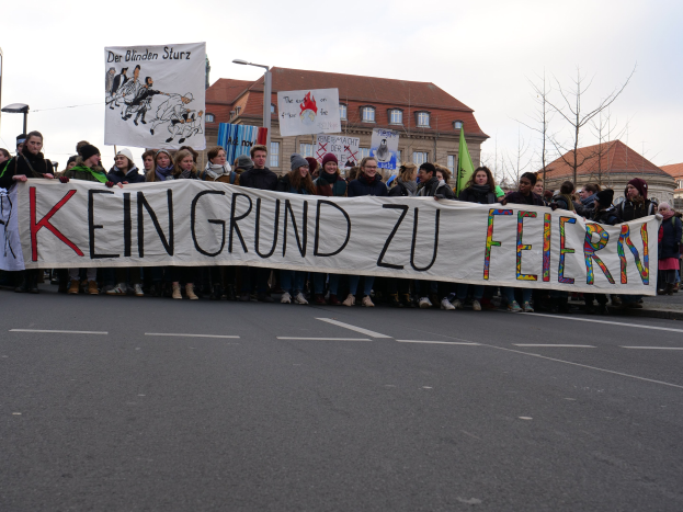 Demonstranten mit einem Transparent "Kein Grund zu Feiern" gegen deutsche Sparmaßnahmen, mit Straßeninfrastruktur und Gebäuden im Hintergrund bei klarem Himmel.