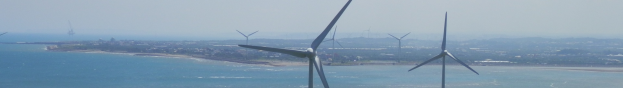 Windkraftanlagen im Meer in der Nähe einer Stadt, mit Wasser, das an den Strand schwappt, Bäumen, Gebäuden und einem klaren blauen Himmel im Hintergrund.