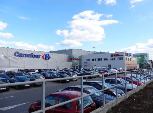 Großes Autohaus mit zahlreichen parkenden Fahrzeugen, umgeben von städtischen Gebäuden, Straßeninfrastruktur und einem bewölkten Himmel.