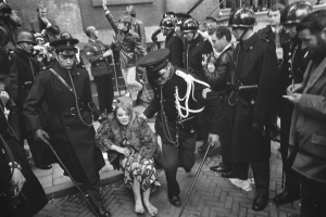 Eine Schwarz-Weiß-Szene einer Frau, die von der Polizei eskortiert wird, vor einer Menge von Menschen mit Helmen und Stöcken, mit einem Gebäude mit Treppe und Geländer im Hintergrund.