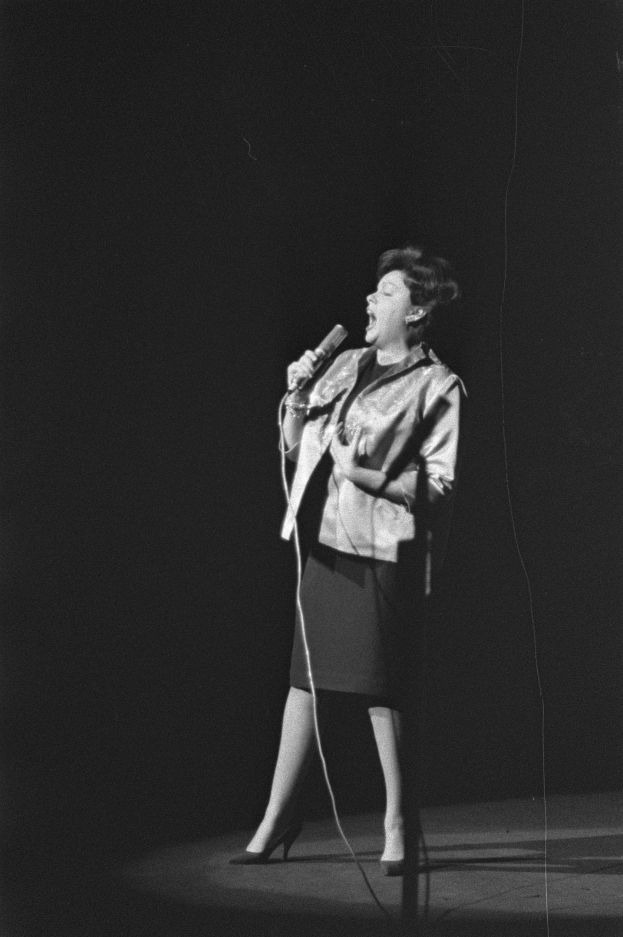 Eine Frau in einem schwarzen und weißen Outfit aus den 1950er Jahren singt fröhlich auf einer von einem Scheinwerfer beleuchteten Bühne und hält ein Mikrofon in der Hand.