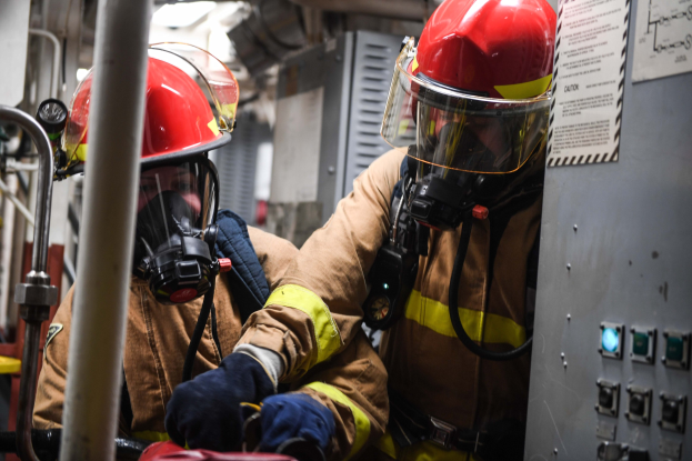 Zwei Feuerwehrleute in Schutzausrüstung, Helmen und Gasmasken bei der Arbeit an einem Feuerwehrauto mit einer Tafel und Text auf der rechten Seite und Metallstangen im Hintergrund.