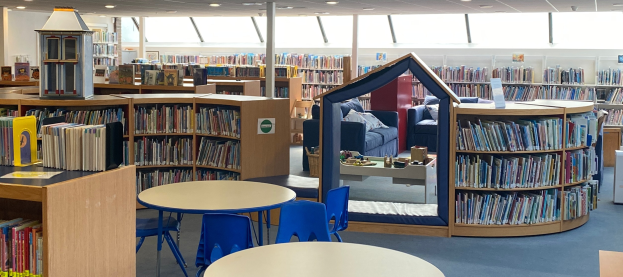 Eine Bibliothek mit Bücherregalen, Tischen und Stühlen, Sofas mit Kissen, einer Säule und Deckenleuchten.