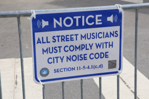 Anschlagtafel an einem Zaun mit einem Schild über Straßenmusiker, die die Stadtlärmvorschriften einhalten, im Hintergrund ist eine Straße zu sehen.
