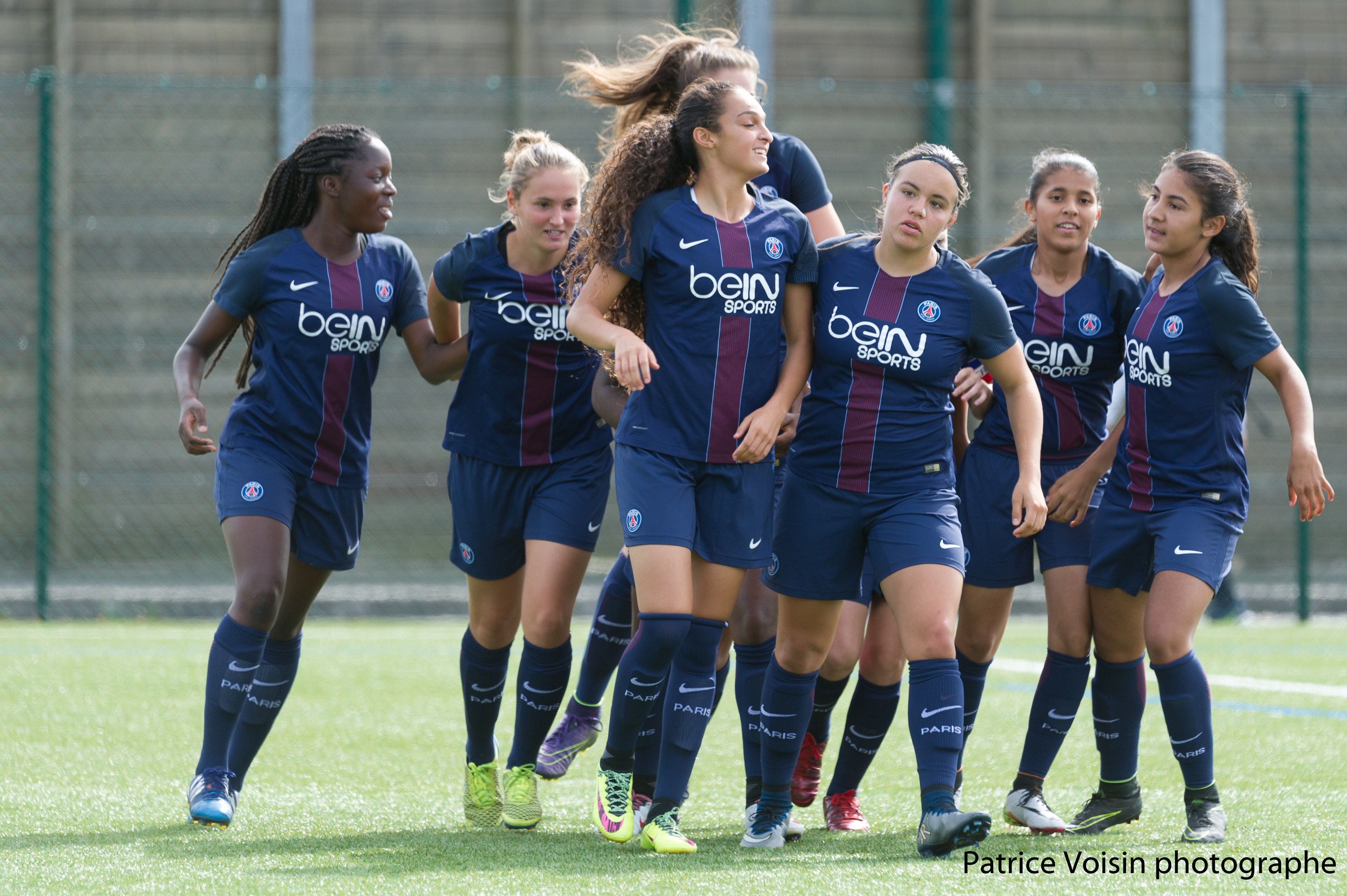 Gruppe junger Frauen beim Fußballspielen auf einem Rasenfeld mit Maschendrahtzaun und einer Wand im Hintergrund, Text in der rechten unteren Ecke lautet "Paris Saint-Germain Frauenfußball".