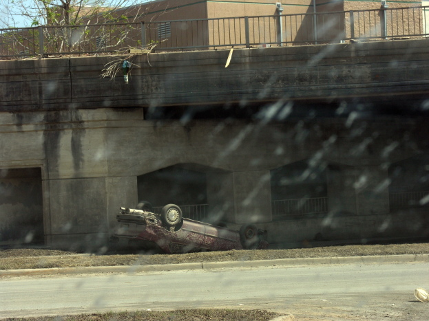 Ein Auto krachte in eine Brücke am Rande einer Straße, umgeben von Gras, mit einem Gebäude mit Gelöndergittern, Bäumen und einem klaren blauen Himmel im Hintergrund.