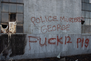 Ein Gebäude mit der Graffiti-Aufschrift "Polizeimord George Fuck 12 P19" an der Seite, das Fenster und verschiedene Gegenstände vorne zeigt.