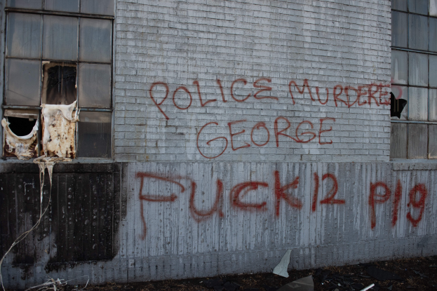 Ein Gebäude mit der Graffiti-Aufschrift "Polizeimord George Fuck 12 P19" an der Seite, das Fenster und verschiedene Gegenstände vorne zeigt.