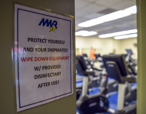 Schriftzug an einer Turnhallentür mit der Aufschrift 'Schützen Sie sich und Ihre Schiffskameraden' mit Laufbändern und Deckenleuchten im Hintergrund.