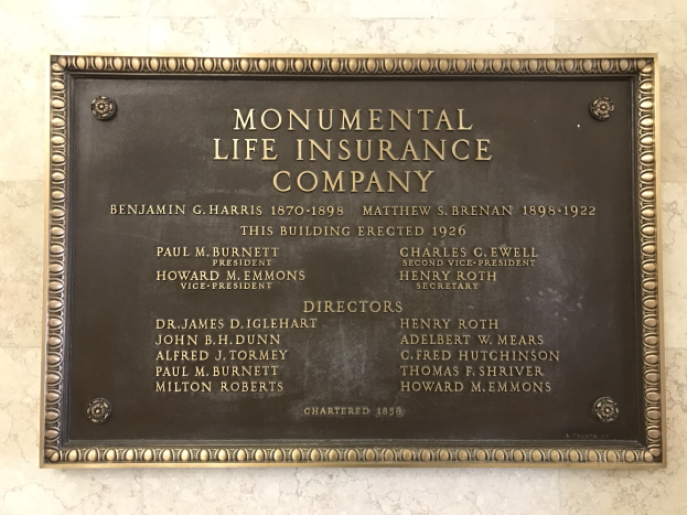 Plakette an einer Gebäudewand mit der Aufschrift "Monumentale Lebensversicherungsgesellschaft" in fetter Schrift, umgeben von einem dekorativen Rahmen auf dunklem Material.