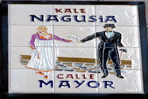 Ein gerahmtes Schild an einem Gebäude trägt die Aufschrift "Kale Nagusia Calle Mayor" mit einem Bild eines Mannes und einer Frau.