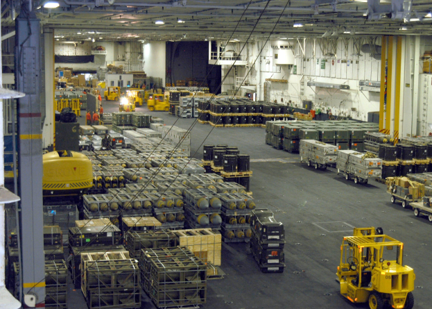 Ein großes Lager mit Lastwagen, Containern, Menschen, Beleuchtungsgeräten, Metallstangen und einem Rad.