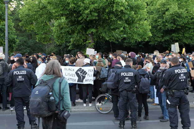 Eine große Gruppe von Menschen nimmt an einer Black Lives Matter Demonstration in Berlin teil, einige halten Schilder und andere tragen Mützen und Taschen, während im Vordergrund ein Fahrrad zu sehen ist und im Hintergrund Bäume und ein Pfahl zu erkennen sind.