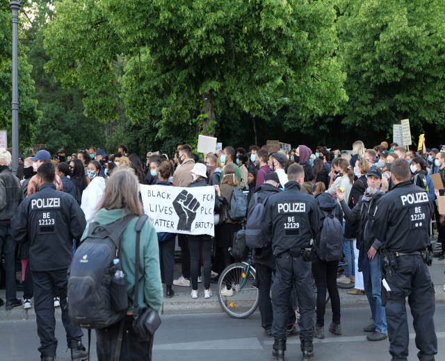 Eine große Gruppe von Menschen nimmt an einer Black Lives Matter Demonstration in Berlin teil, einige halten Schilder und andere tragen Mützen und Taschen, während im Vordergrund ein Fahrrad zu sehen ist und im Hintergrund Bäume und ein Pfahl zu erkennen sind.