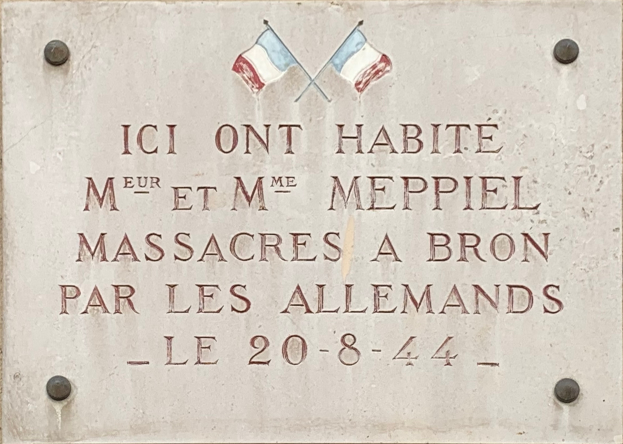 Ein Steintafel an einem Gebäude mit zwei Flaggen, beschriftet mit Text, der ein Massaker gedenkt.