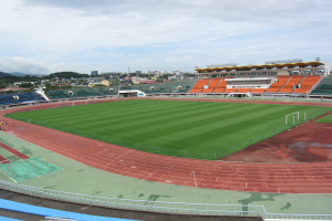Ein großes Stadion mit einem Fußballfeld in der Mitte, umgeben von Gebäuden, Bäumen und Hügeln unter einem klaren blauen Himmel, mit wenigen Menschen und saftigem grünem Gras auf dem Boden.