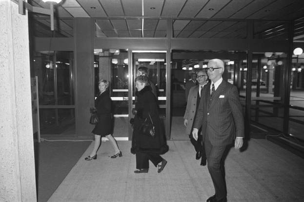 Eine Gruppe von Menschen in Anzügen geht auf einem Gehweg vor einem Gebäude vorbei, das von Deckenlampen beleuchtet wird, auf einem Schwarz-Weiß-Bild.