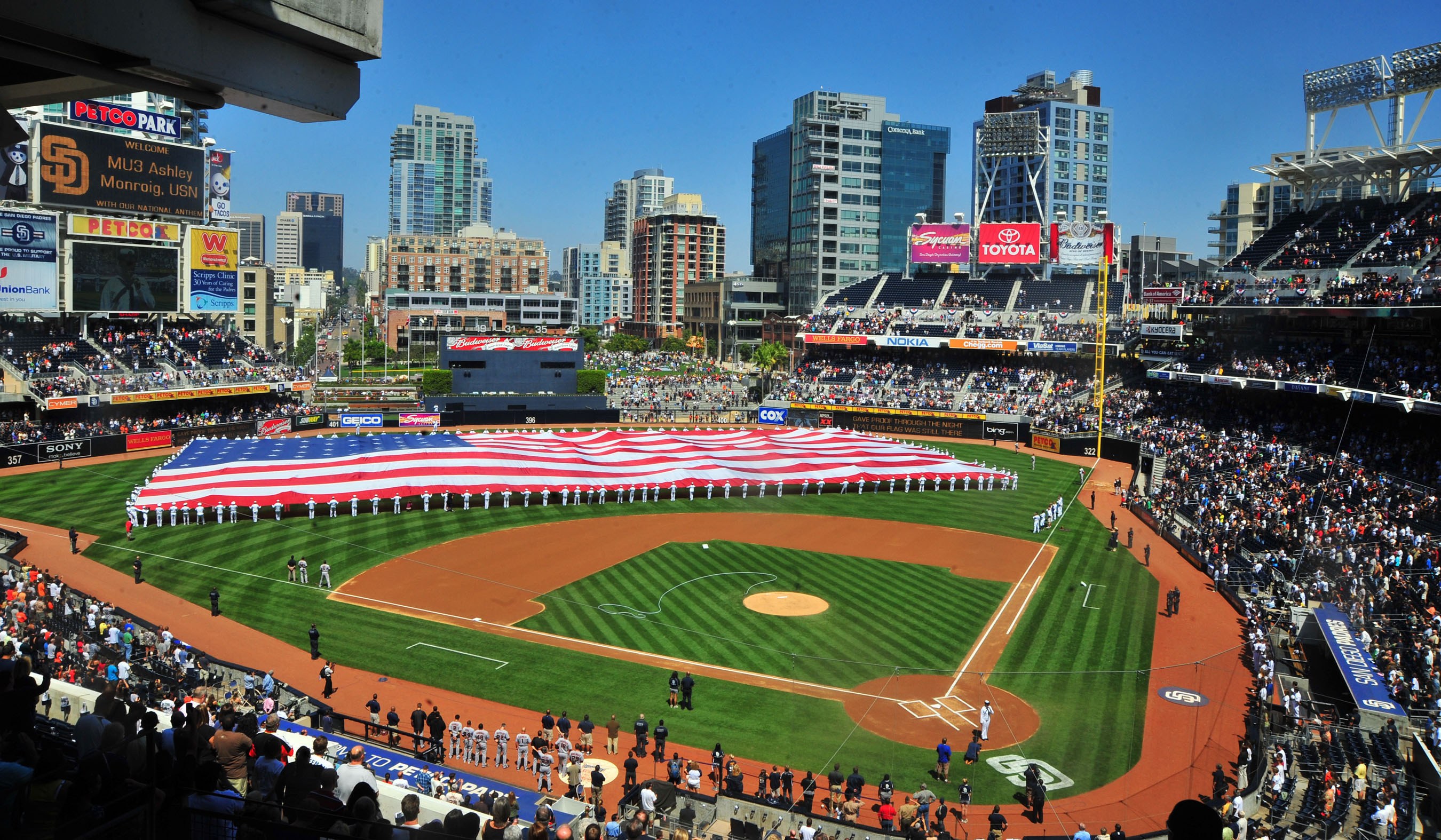 Baseballstadion voller Zuschauer, mit einer Gruppe unter einer amerikanischen Flagge auf dem Feld, Anzeigetafeln auf beiden Seiten und Gebäuden, Bäumen und einem klaren Himmel im Hintergrund.