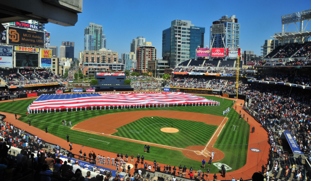 Baseballstadion voller Zuschauer, mit einer Gruppe unter einer amerikanischen Flagge auf dem Feld, Anzeigetafeln auf beiden Seiten und Gebäuden, Bäumen und einem klaren Himmel im Hintergrund.