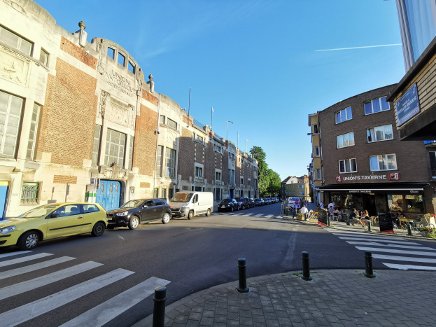 Eine Stadtstraße mit parkenden Autos, Gebäude mit Fenstern, Menschen, die gehen, eine Zebrastreifen in der Nähe, Bäume und einen klaren blauen Himmel im Hintergrund.