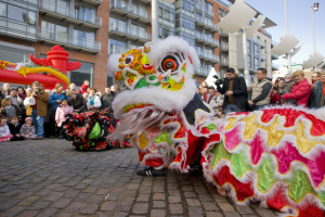 Ein lebendiges chinesisches Neujahrsfest in Amsterdam mit einer Löwen-Tanz-Show vor einer Zuschauermenge, darunter einige, die das Ereignis fotografieren, vor einem Gebäudehintergrund und einem klaren blauen Himmel.
