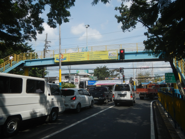 Eine belebte Straße mit Fahrzeugen, eine Brücke mit Geländern und Treppen, Laternen, Verkehrszeichen, Informationsschilder, Bäume, Gebäude und ein bewölkter Himmel.