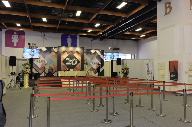 Großer Ausstellungssaal mit Tischen und Stühlen in Reihen, Absperrpoller mit Bändern, Blumenbouquets, Rednerpulte, Banner, einer Wand mit dem Buchstaben B und Deckenleuchten.