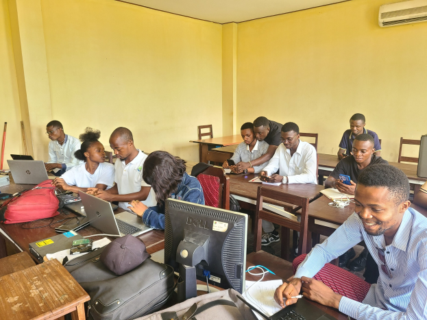Eine Gruppe von Menschen sitzt an Tischen in einem Klassenzimmer mit Laptops, Taschen, Monitoren und Büchern auf den Tischen und einer Klimaanlage an der Wand im Hintergrund.
