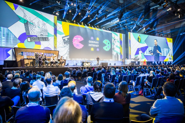 Eine große Menschenmenge, die auf Stühlen vor einer Bühne sitzt, mit einigen Personen auf dem Podium im Vordergrund, einem Tisch mit verschiedenen Gegenständen links und Bildschirmen und Lichtern im Hintergrund auf der Deutschen Internet-Konferenz 2019.