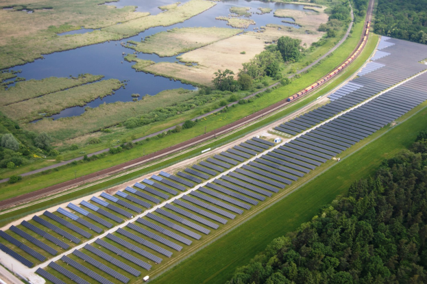 Luftaufnahme einer Solaränlage mit Panelen, umgeben von Bäumen, Gras, Wasser und einer nahen Schiene.