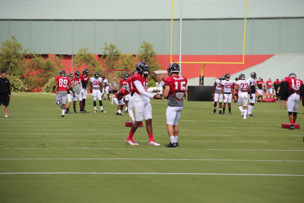 Gruppe von Menschen, die Fußball auf einem Rasenfeld mit Helmen und Fußballschuhen spielen, umgeben von Bäumen, Pfosten, einer Wand und einem klaren blauen Himmel.