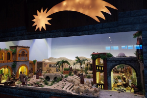 Eine Krippe Ausstellung im Museum, mit Menschen, Tieren, Häusern, Bäumen und anderen Gegenständen, beleuchtet von einer Deckenbeleuchtung vor einer Wand.