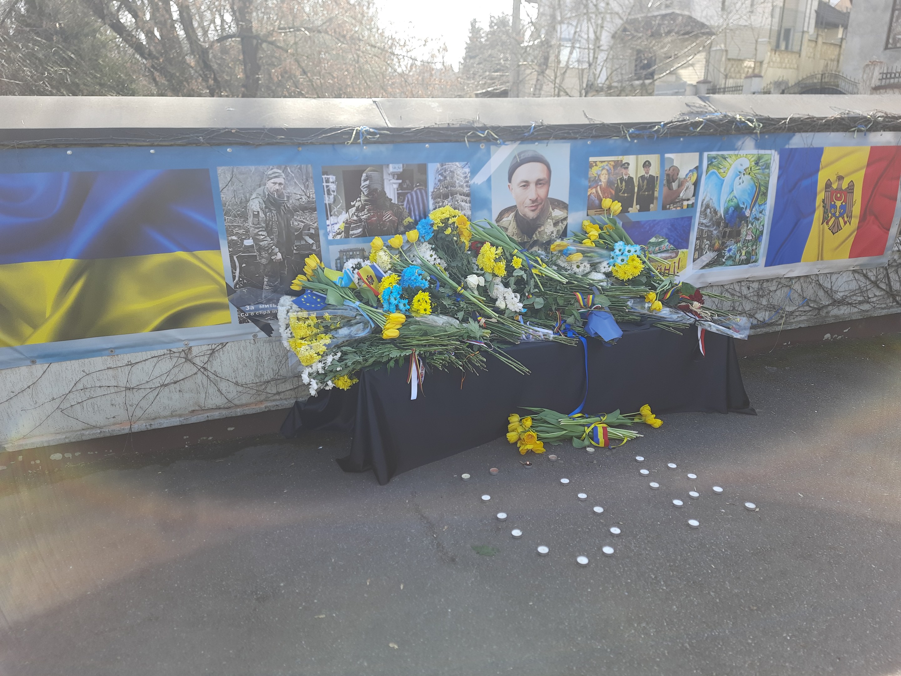 Ein provisorisches Denkmal in Kiew, Ukraine, mit Blumensträußen auf der Straße, Fotos und Flaggen an einer Wand, Bäumen, Gebäuden und einem klaren blauen Himmel im Hintergrund.