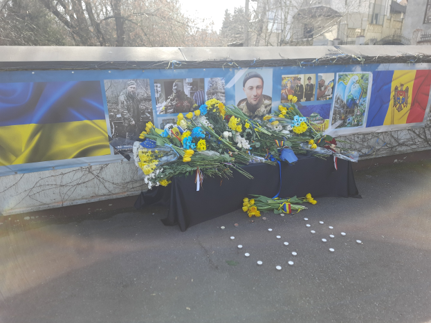 Ein provisorisches Denkmal in Kiew, Ukraine, mit Blumensträußen auf der Straße, Fotos und Flaggen an einer Wand, Bäumen, Gebäuden und einem klaren blauen Himmel im Hintergrund.