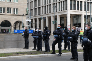 Eine Gruppe von Polizisten in schwarzen Uniformen und Masken steht vor einem Gebäude mit Glasfenstern und Säulen in Berlin, Deutschland, einige halten Helme, mit Laternen, Verkehrszeichen, Schildern mit Text, einer Statue, anderen Menschen und grasbewachsenem Boden im Hintergrund.