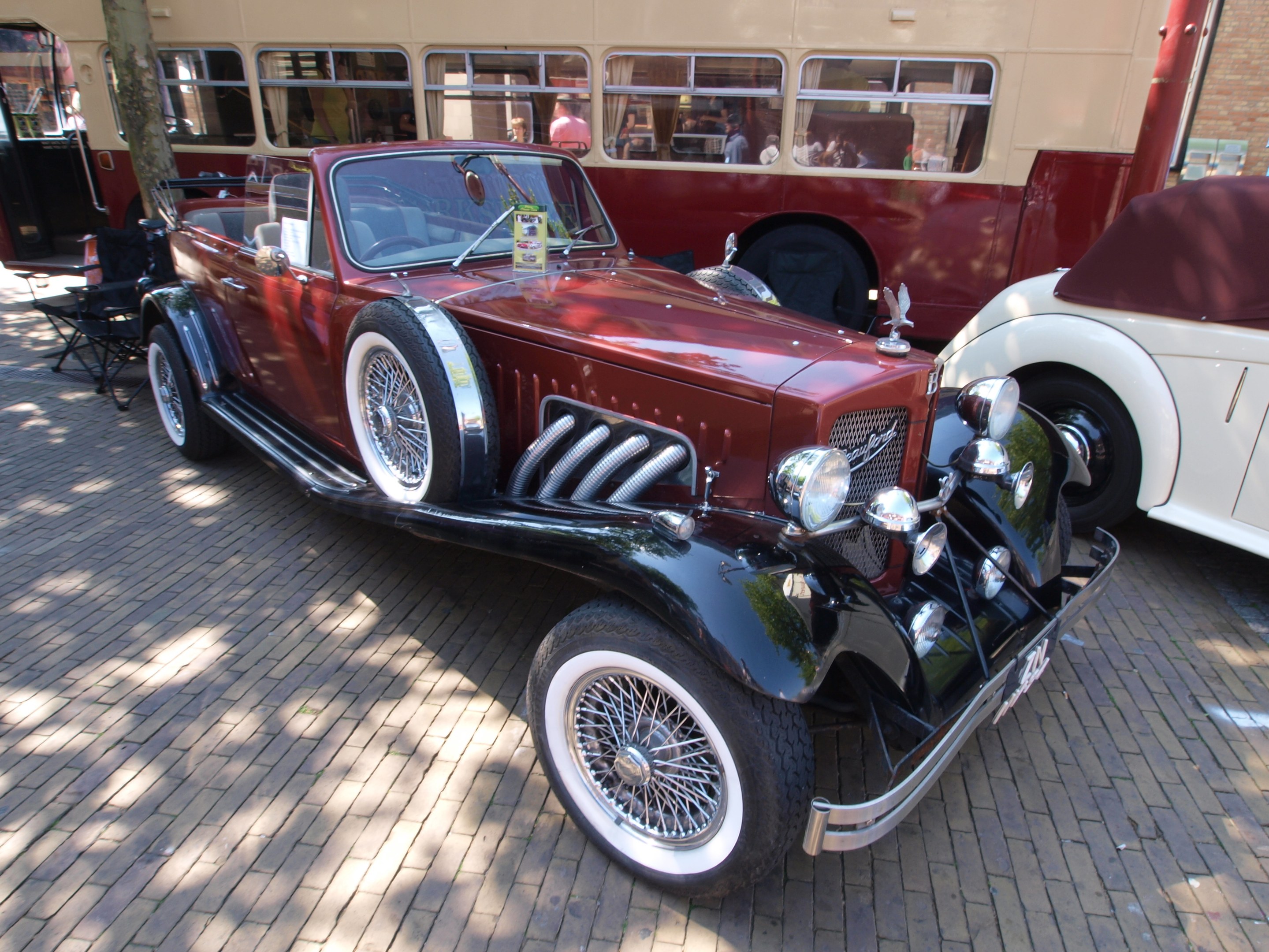 Ein Oldtimer neben einem Doppeldeckerbus auf einer Kopfsteinpflasterstraße, mit Stühlen und einem Baum im Vordergrund, einem Gebäude mit Fenstern im Hintergrund und Menschen im Bus zu sehen.