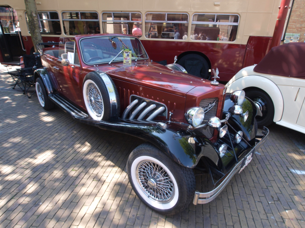 Ein Oldtimer neben einem Doppeldeckerbus auf einer Kopfsteinpflasterstraße, mit Stühlen und einem Baum im Vordergrund, einem Gebäude mit Fenstern im Hintergrund und Menschen im Bus zu sehen.