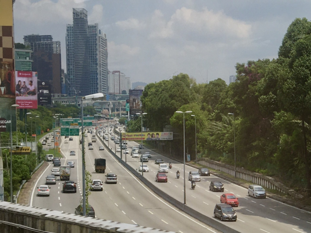 Eine vielbefahrene Autobahn mit hohen Gebäuden, Straßenlaternen, Werbetafeln, Bäumen und einem bewölkten Himmel.