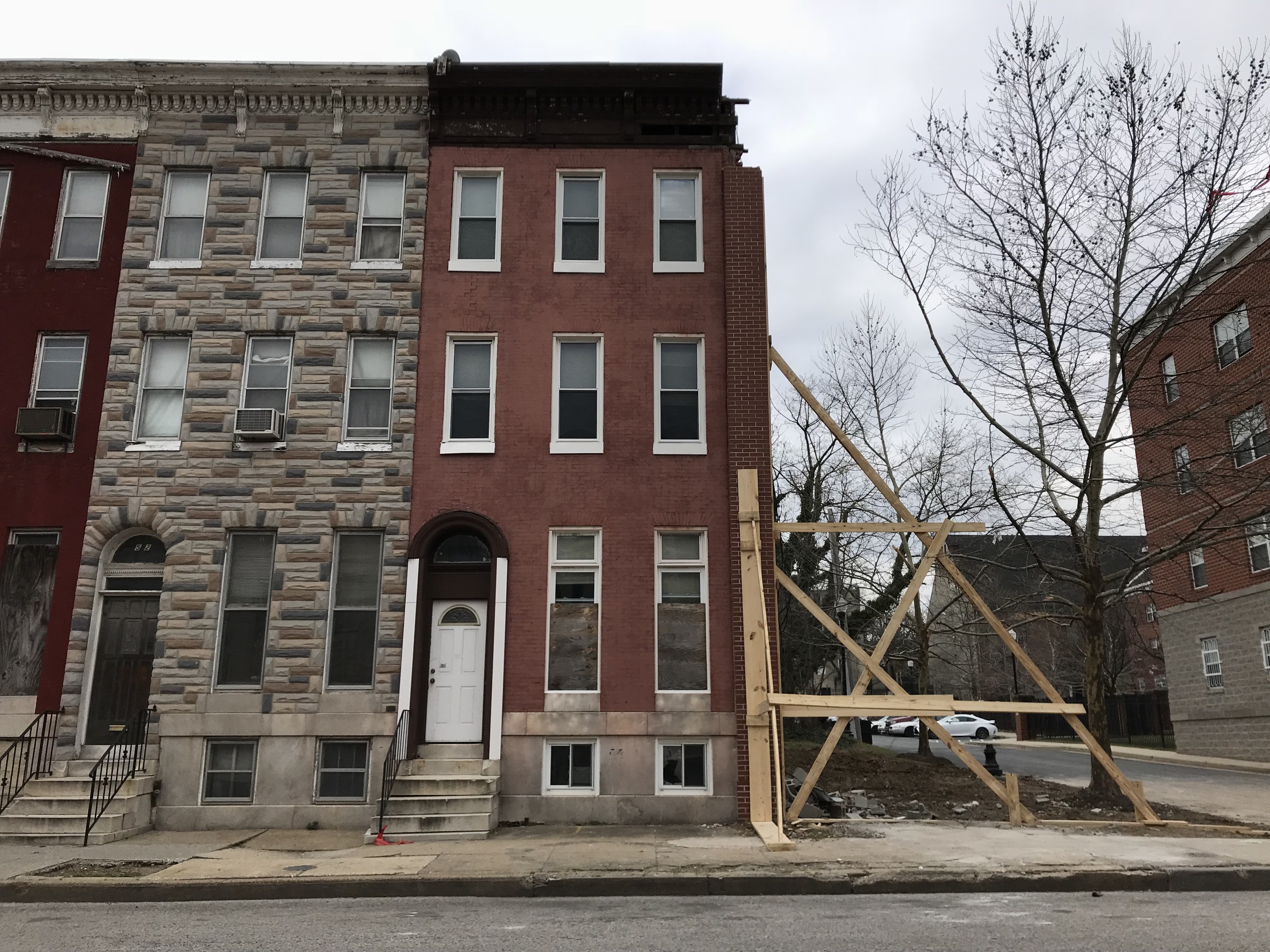 Eine Reihe von Häusern an einer Straßenecke mit sichtbaren Renovierungen, umgeben von verstreuten Werkzeugen und Materialien, Bäumen, Fahrzeugen und einem klaren blauen Himmel.