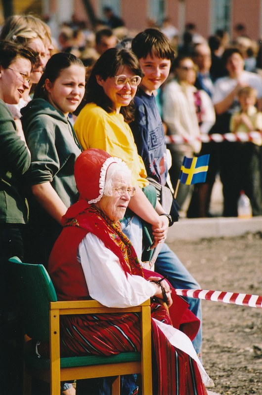 Alte Frau sitzend in einem Stuhl vor einer Menge, mit einer Schleife im Vordergrund und einem Gebäude im Hintergrund.