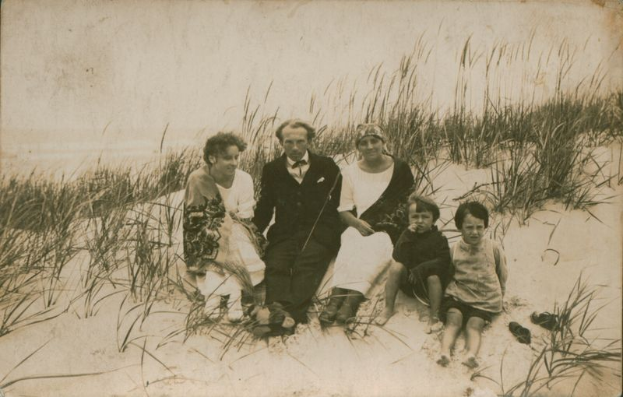 Ein altes Schwarz-Weiß-Foto einer Familie, die auf einem Strand sitzt, umgeben von Sand und Gras.