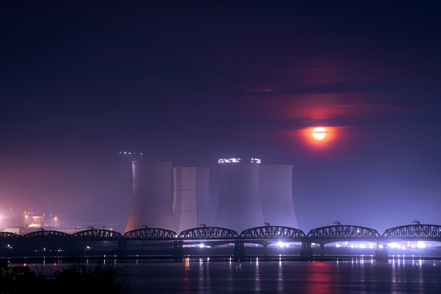Ein Kernkraftwerk bei Nacht, mit einer Brücke über einen Gewässer, beleuchtet von Gebäuden und Mondlicht, umgeben von Bäumen.