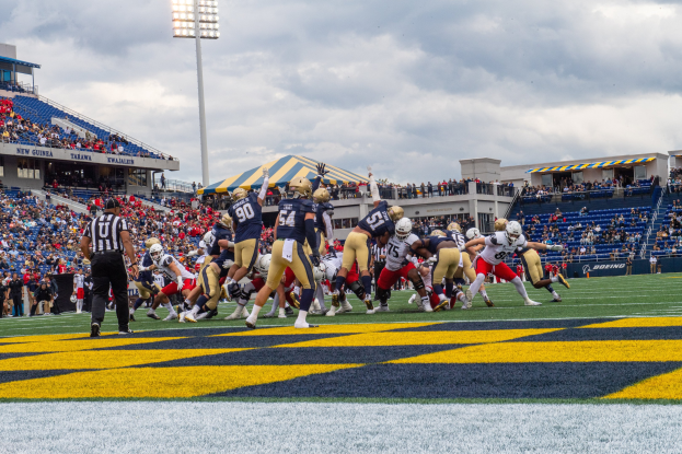 Fußballspieler mit Helmen und Sportkleidung auf einem Feld mit Zuschauern im Hintergrund, Stadionstrukturen und Text unten.